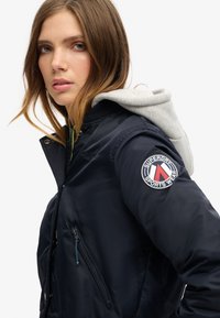 Marineblå jakke med grå hætte, der har et logo-patch på ærmet, lynlås-lommer og en glat, blank overflade.