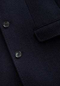 Abrigo de lana azul marino con una textura especial, dos botones negros y un detalle de bolsillo cosido. La vista en detalle captura el material y color.