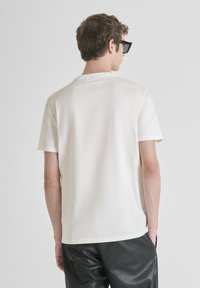 Camiseta blanca de algodón con mangas cortas, escote redondo y un sutil logo en la parte posterior; modelo lleva pantalones de cuero negros y gafas de sol.