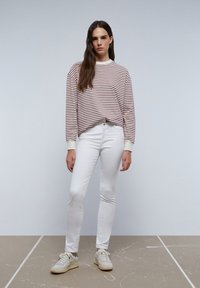 Gestreepte, oversized lang mouw shirt in crèmekleur en bordeaux; gecombineerd met witte slim-fit jeans en lichtkleurige sneakers. Het geheel heeft een soepele textuur.