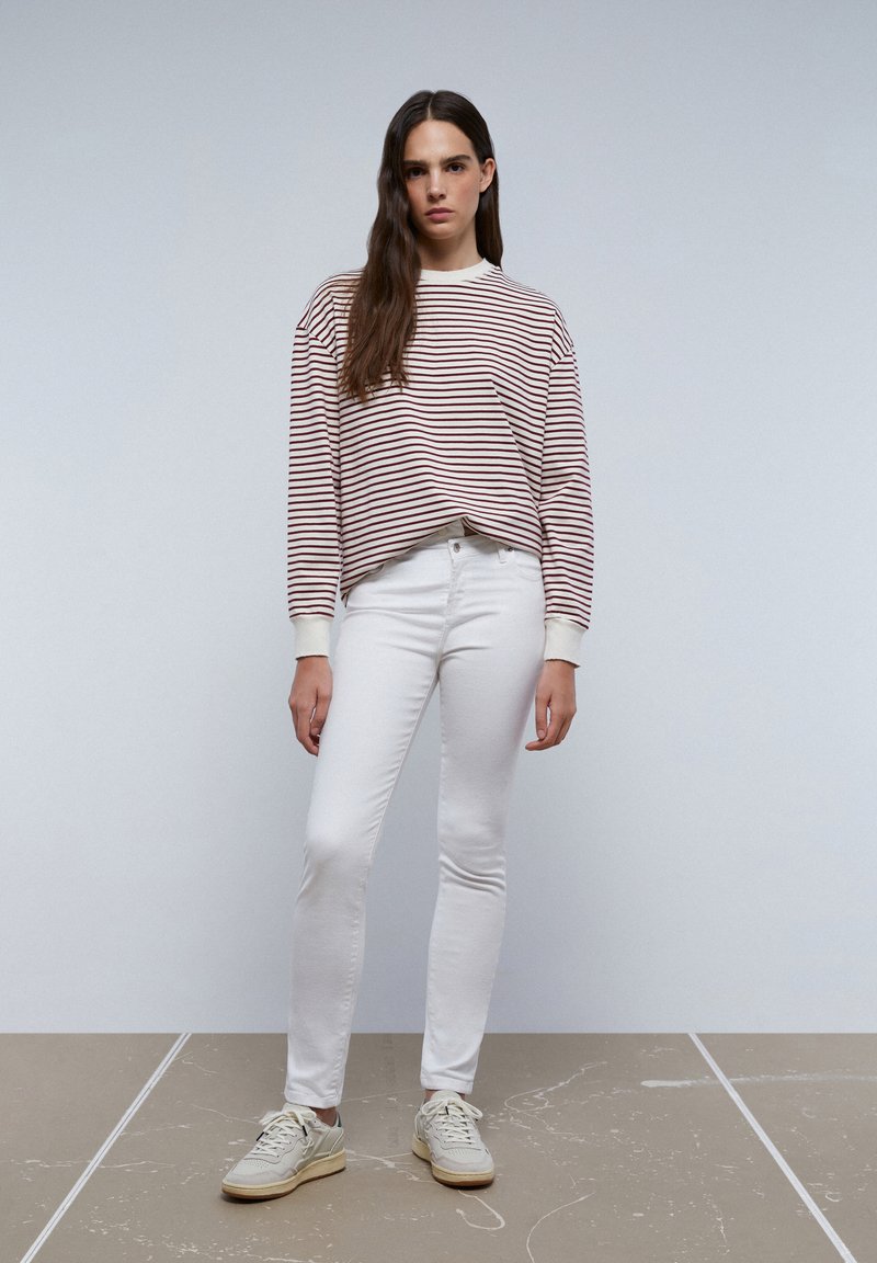 Gestreepte, oversized lang mouw shirt in crèmekleur en bordeaux; gecombineerd met witte slim-fit jeans en lichtkleurige sneakers. Het geheel heeft een soepele textuur.