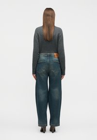 Γυναίκα με ίσια καστανά μαλλιά, που φοράει γκρι πουλόβερ, ξεθωριασμένο τζιν φαρδιάς γραμμής και καφέ παπούτσια με τακούνι, εμφανιζόμενη από την πίσω πλευρά.