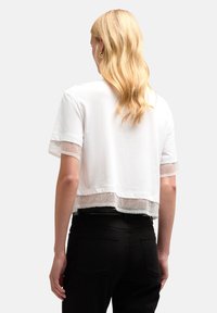 Camiseta blanca recortada con mangas transparentes y texturizadas. Presenta un dobladillo recto y una tela suave. Se lleva con pantalones negros.