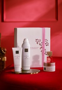 Gavesettet inkluderer tre hvite og rosa Rituals-produkter: kroppskrem, skummende dusjgelé og kroppsskrubb, på en gullfat mot en rød bakgrunn.