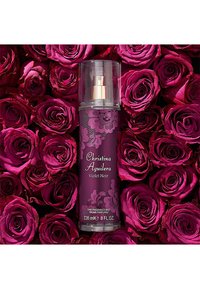 Bottiglia di spray profumato viola con design floreale e accenti dorati, posizionata tra vivaci rose rosa. Dimensione 236 mL.