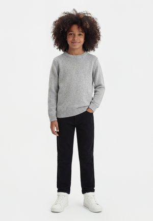 Enfant aux cheveux bouclés portant un pull gris, un pantalon noir et des baskets blanches, debout avec une main dans la poche sur fond blanc.