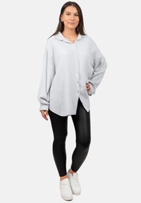 Camicia button-up grigia chiara con texture, vestibilità ampia, maniche oversize e orlo curvo; abbinata a leggings neri e sneakers bianche.