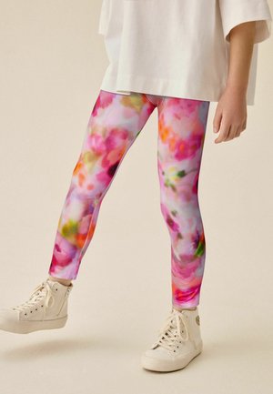 Kind draagt witte hoge sneakers en kleurrijke roze, oranje en groene gebloemde legging met een los wit shirt.
