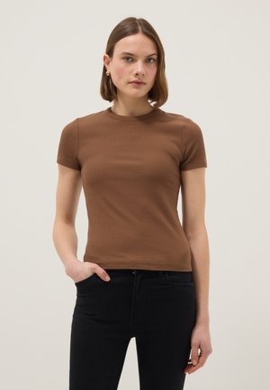 Frau mit kurzen braunen Haaren, die goldene Ohrringe, ein eng anliegendes braunes Kurzarmoberteil und schwarze Hose trägt, steht mit einer Hand in der Tasche.