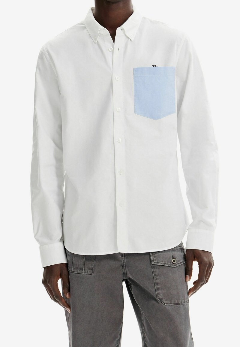 Homme portant une chemise blanche à boutons avec une poche poitrine bleu clair et un pantalon cargo gris, debout devant un fond blanc.