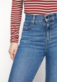 Jeans de cintura alta de color azul con un lavado desgastado, bolsillos frontales y un botón plateado, combinados con una camiseta de manga larga a rayas rojas y blancas.