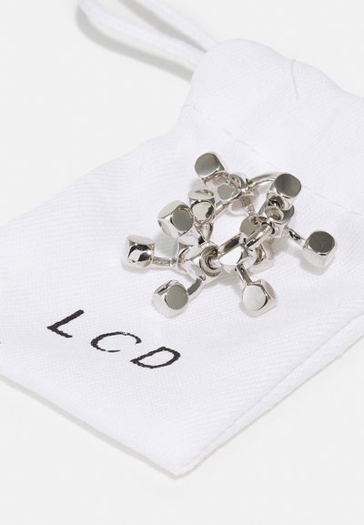 Pièce de bijou en métal argenté avec un design géométrique, posée sur un sachet en coton blanc portant l'inscription "LCD" imprimée en noir.