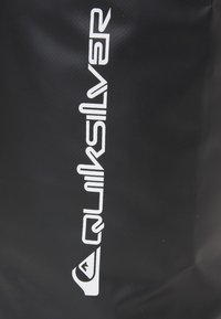 Bolsa negra con un logo blanco prominente. El material es texturizado y el diseño es elegante con una apariencia minimalista.