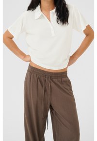 Jasna kremowa koszulka polo z kołnierzykiem i trzema guzikami, w połączeniu z luźnymi, brązowymi spodniami na gumce. Prosty, casualowy design.
