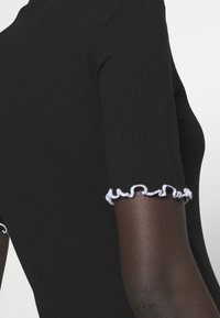 Top negro de manga corta acanalado con bordes rizados blancos, llevado sobre un brazo de piel oscura contra un fondo claro y liso.