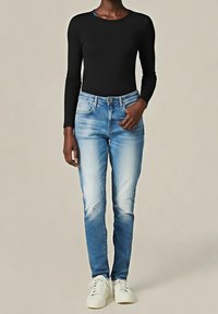 Personne portant un jean bleu clair délavé, un t-shirt noir à manches longues et des baskets blanches, debout avec une main dans la poche.