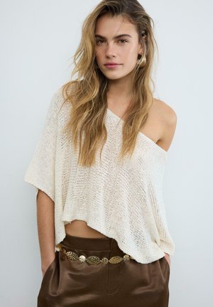 Mladá žena s dlhými blond vlasmi nosí pohodlný krémový sveter so spustenými ramenami, hnedé kožené nohavice a zlatý ozdobný opasok, stojí so založenými rukami vreckách.