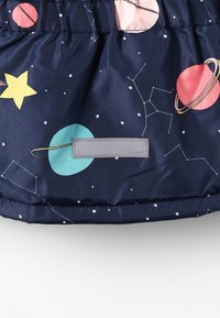 Tissu bleu marine avec des planètes colorées, des étoiles et des motifs de constellations, comportant une petite étiquette réfléchissante rectangulaire cousue près du bas.