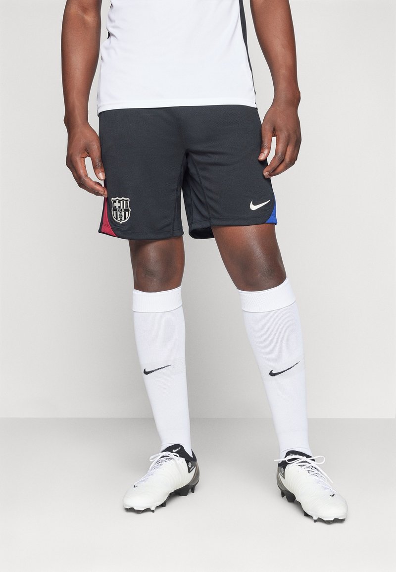 Homme portant un short de football noir du FC Barcelone, des chaussettes blanches Nike et des chaussures de football blanches, debout devant un fond uni.