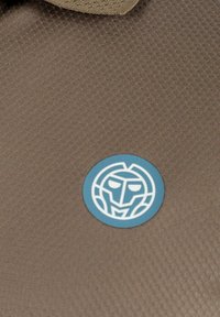 Ein brauner, strukturierter Stoff zeigt einen auffälligen blauen, kreisförmigen Aufnäher mit einem stilisierten weißen Gesichtsmuster.