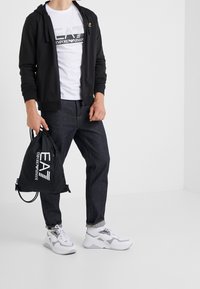 Svart zip-up hoodie, vit t-shirt med logga, mörka jeans, vita och grå sneakers. Håller en svart dragsko-väska med logga.