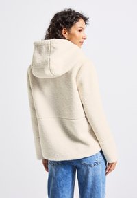 Femme avec des cheveux bouclés portant un hoodie en polaire crème et un jean bleu, légèrement tournée sur le côté devant un fond uni.