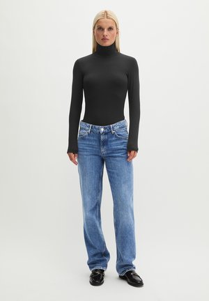 Blonde Frau steht und trägt ein schwarzes, langärmeliges Rollkragentop, blaue hoch taillierte Jeans und schwarze Schuhe vor einem einfarbigen Hintergrund.