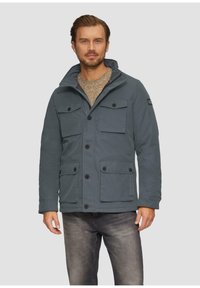 Graue Jacke mit hohem Kragen und Knopfverschluss, die mehrere Fronttaschen und einen strukturierten Pullover darunter hat.