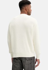 Maglione lavorato a maglia color crema con motivo testurizzato, polsini e orlo a coste, e vestibilità rilassata, indossato con jeans grigio scuro.