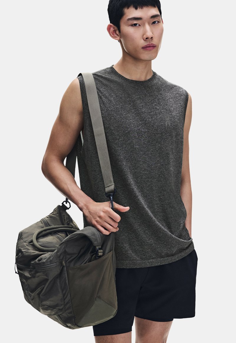 Jeune homme portant un débardeur gris et un short noir, portant un grand sac de sport vert olive sur une épaule.