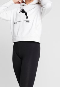 Persona che indossa un hoodie bianco della Puma con logo nero e leggings neri, mentre sistema il cappuccio su uno sfondo chiaro e semplice.