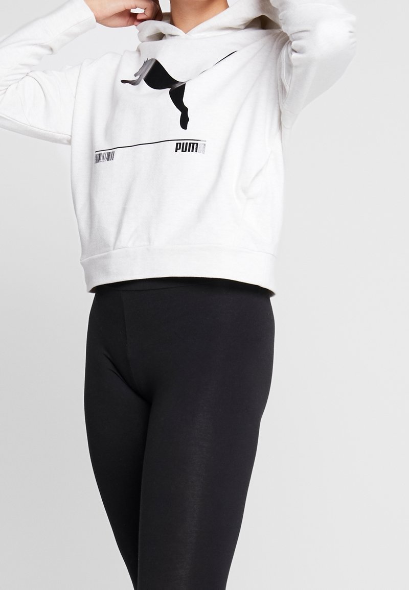 Persona che indossa un hoodie bianco della Puma con logo nero e leggings neri, mentre sistema il cappuccio su uno sfondo chiaro e semplice.