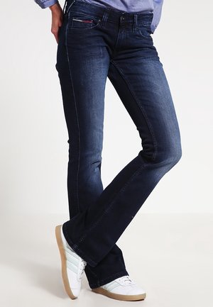 Osoba nosząca ciemnoniebieskie obcisłe jeansy bootcut i białe trampki z jasnoniebieskimi paskami, stojąca ze skrzyżowanymi nogami.