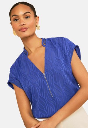 Kobieta w niebieskim, teksturowanym topie z krótkim rękawem i suwakiem z przodu oraz złotych kolczykach w kształcie liści, pozująca na jednolitym jasnym tle.