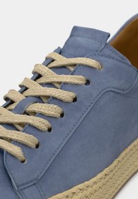 Μπλε σουέντ αθλητικό παπούτσι με πλεγμένα μπεζ κορδόνια και σόλα από τζούτ. Διαθέτει στρογγυλεμένη μύτη και ελάχιστες ραφές στο πάνω μέρος.