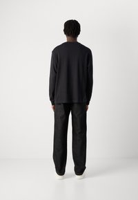 Mann med korte dreadlocks står med ryggen til iført en svart langermet skjorte, mørke bukser og hvite sneakers mot en ensfarget bakgrunn.