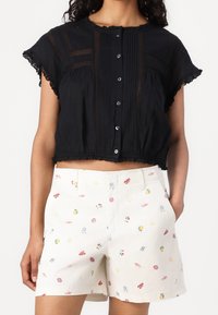 Top corto nero con maniche arricciate, chiusura frontale con bottoni e dettagli testurizzati. Abbinato a pantaloni corti crema con motivi ricamati colorati.