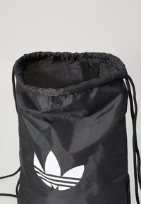 Saco de cordão preto feito de nylon brilhante com uma textura suave, apresentando um grande logótipo branco de um lado e alças de corda ajustáveis.
