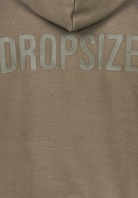 Brun hætte sweatshirt med stort gråt tekst, der står "DROPSIZE" på ryggen. Materialet ser blødt ud med en glat tekstur.