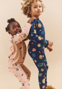 Twee kinderen in bijpassende pyjama's. Een heeft een lichtroze top met kleine zwarte bladeren, de ander een blauwe top met kleurrijke planeten en zonnen.