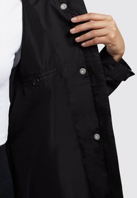 Veste noire avec une finition lisse et mate. Équipée de boutons-pression et d'un détail de poche subtil. Le tissu semble léger et flexible.