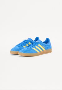 Blå ruskindsneakers med gule tre-striber og guld bogstaver. Udstyret med en gummisål og en lav profil.