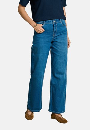 Person, die weite blaue Jeans, braune flache Schuhe und ein dunkles Oberteil trägt, mit einer Hand in der Tasche vor einem schlichten Hintergrund.