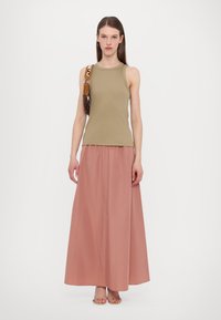 Top senza maniche verde oliva abbinato a una lunga gonna rosa fluida; il modello tiene una borsa con una tracolla a fantasia. Texture liscia e vestibilità rilassata.