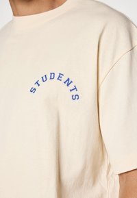 Beige bomull t-skjorte med blå brodert "STUDENTS" i en bue-design på brystet. Løs passform med korte ermer.