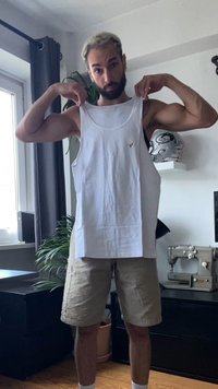 Top in cotone bianco sollevato, con scollo rotondo e un piccolo logo dorato. Il modello indossa pantaloni beige, in piedi all'interno con piante attorno.