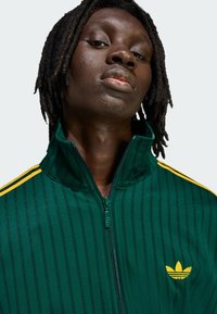 Zelená bunda na zips z hladkej látky s vertikálnymi pruhmi a žltými akcentmi. Na ľavej strane na hrudi je zobrazené ikonické logo.