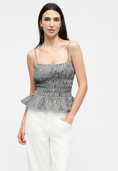 Γυναίκα με μακριά σκούρα μαλλιά που φορά κρίκους σκουλαρίκια, ριγέ ασπρόμαυρο ρούχο με λεπτές τιράντες και λευκό ψηλόμεσο παντελόνι με το χέρι στην τσέπη.