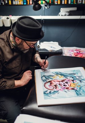 Tatér v černém klobouku kreslí ilustraci opice s korunou, obklopené tropickým listím, na bílém papíře.