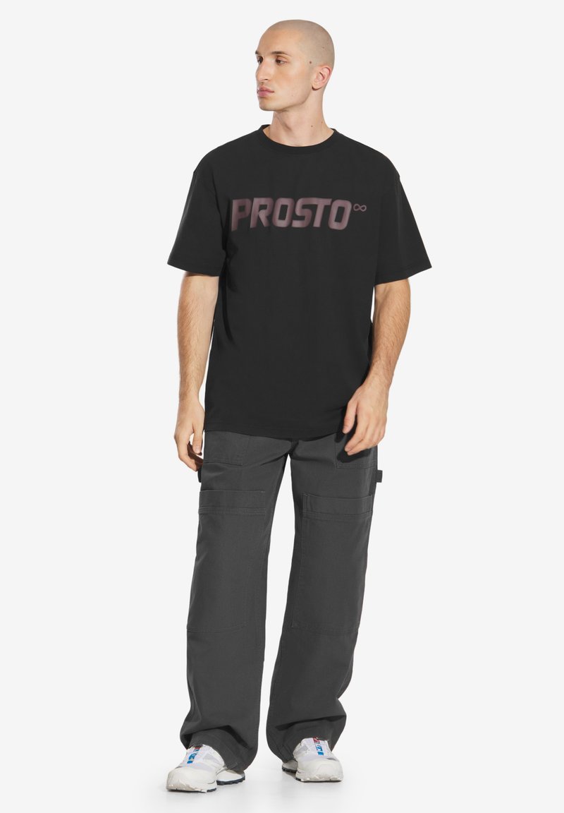 Czarna koszulka z okrągłym dekoltem z dużą grafiką "PROSTO" w metalicznym odcieniu; stylizowana z ciemnoszarymi spodniami cargo i białymi trampkami.